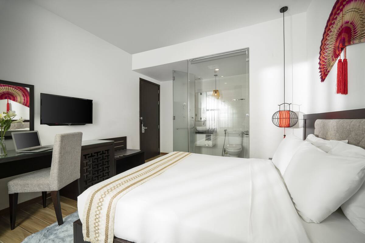 A modern hotel room with a bed, desk, chair, TV, and a glass shower area.