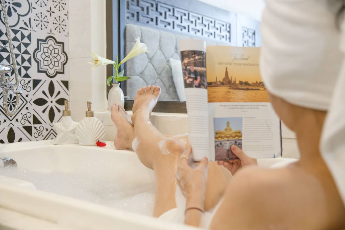 A person relaxing in a bathtub with a book and a vase of flowers.