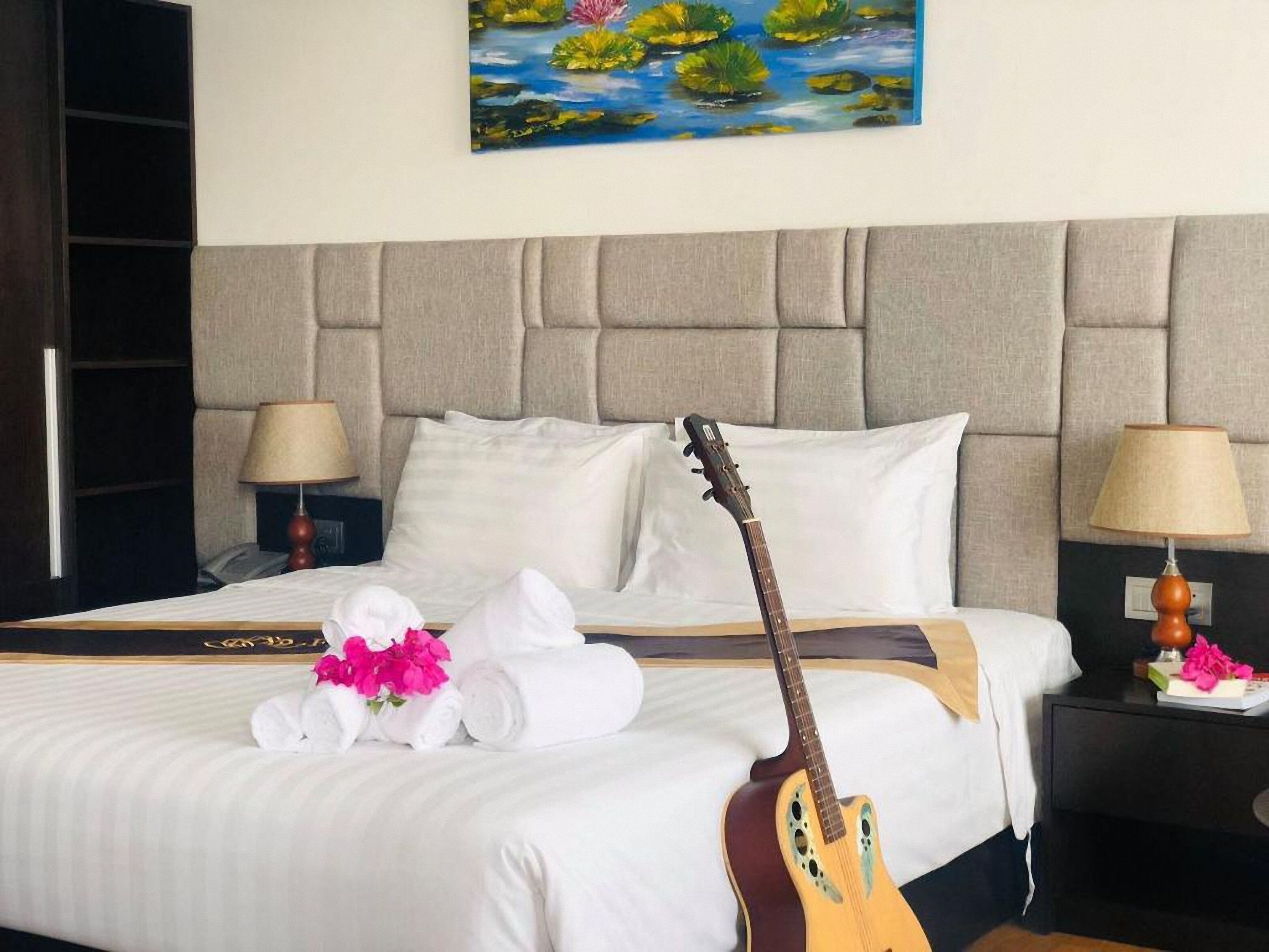 A hotel room with a bed, pillows, a guitar, towels, and a painting of a pond with flowers.