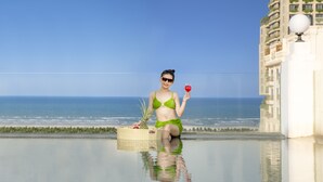 Une personne vêtue d’un bikini vert est assise au bord d’une piscine, avec un panier en osier et une boisson rouge à proximité. Elle a devant elle une plage et l’océan.