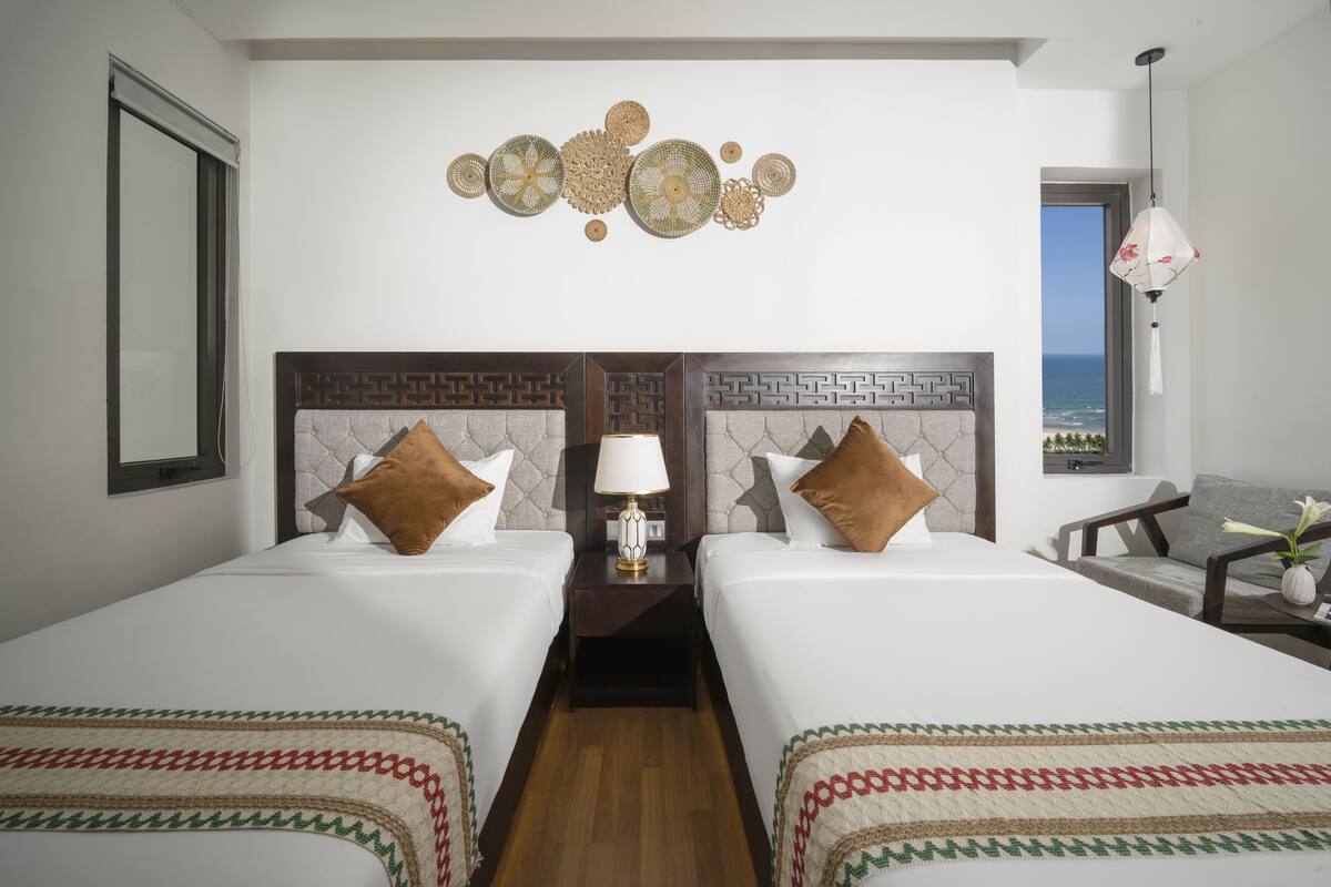 A hotel room with two beds, a nightstand with a lamp, a decorative wall hanging, and a view of the ocean.