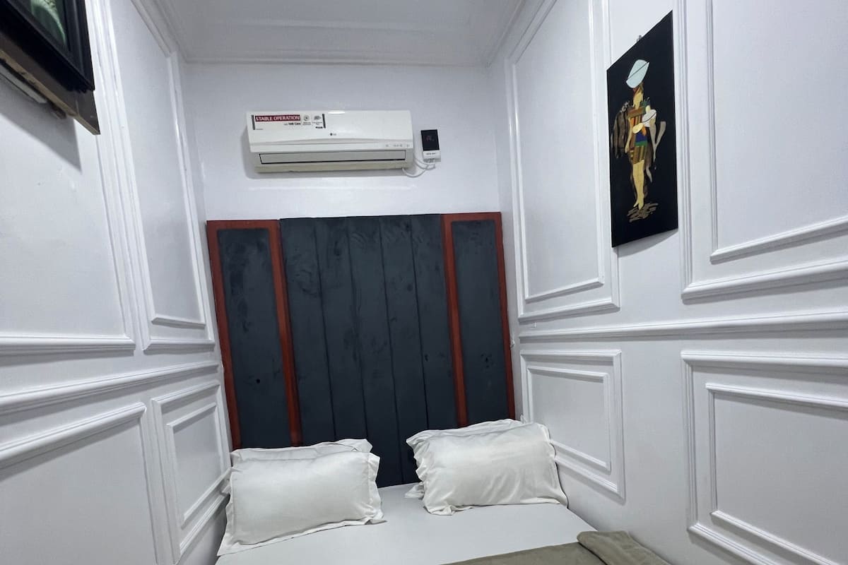 A small, neatly arranged bedroom with a bed, pillows, an air conditioning unit, and a framed picture on the wall.