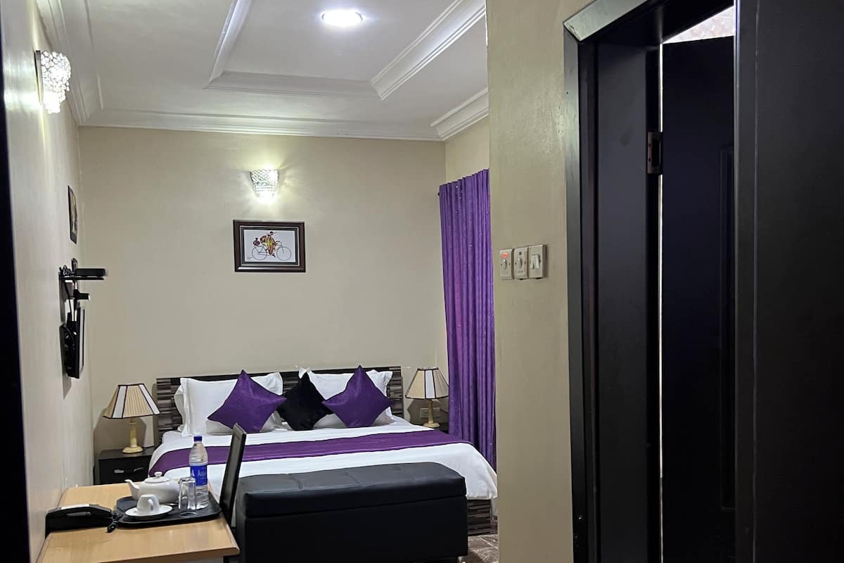 A hotel room with a bed, a desk with a chair, a nightstand, a lamp, and a bathroom door.