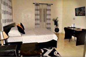 A hotel room with a bed, a desk, a chair, a lamp, a plant, and a TV. - J. Gibson Hotel (Lekki)