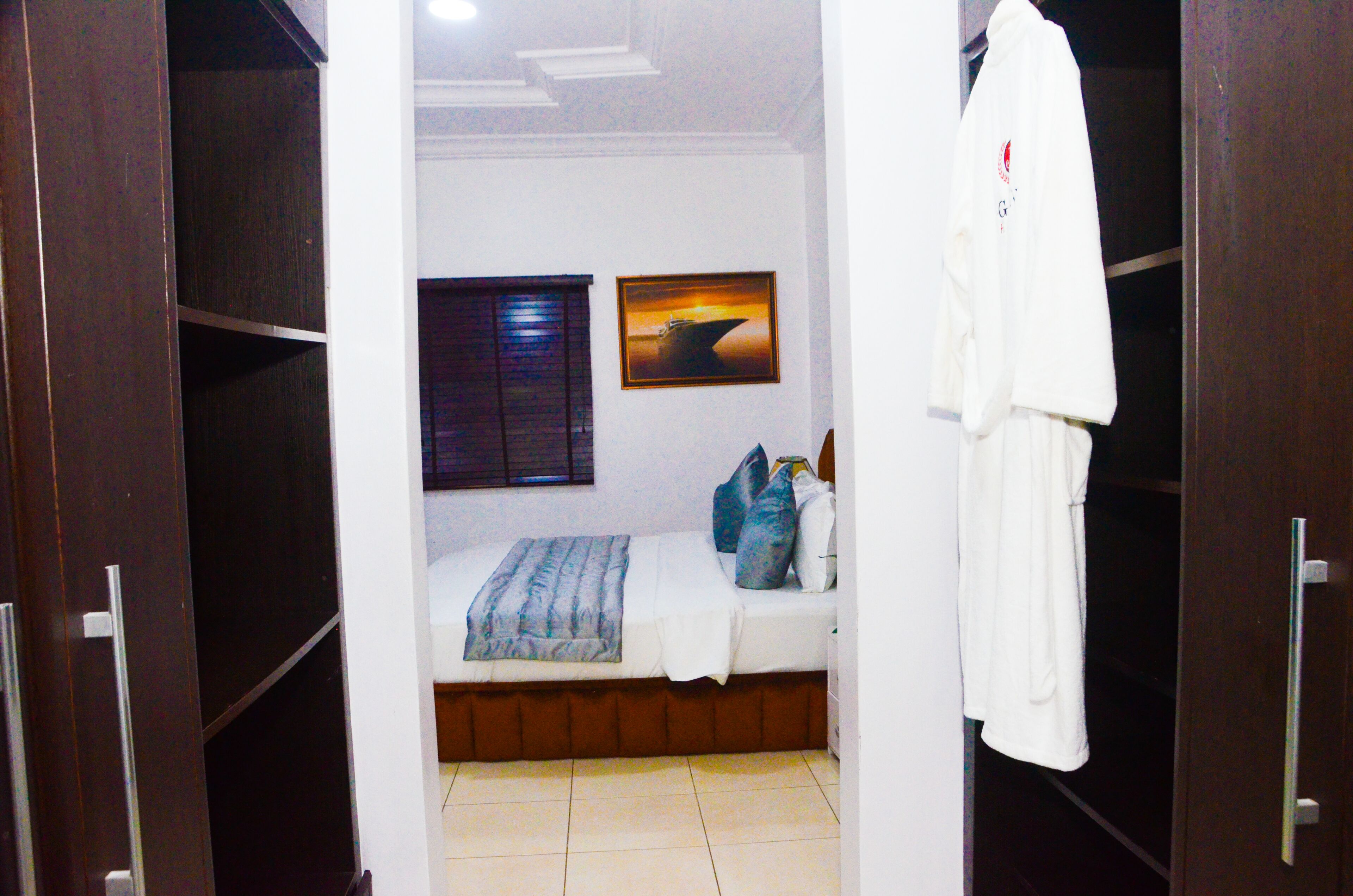A bedroom with a bed, pillows, a blanket, a window with blinds, a framed picture on the wall, and a robe hanging on a door.