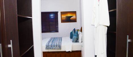 A bedroom with a bed, pillows, a blanket, a window with blinds, a framed picture on the wall, and a robe hanging on a door.