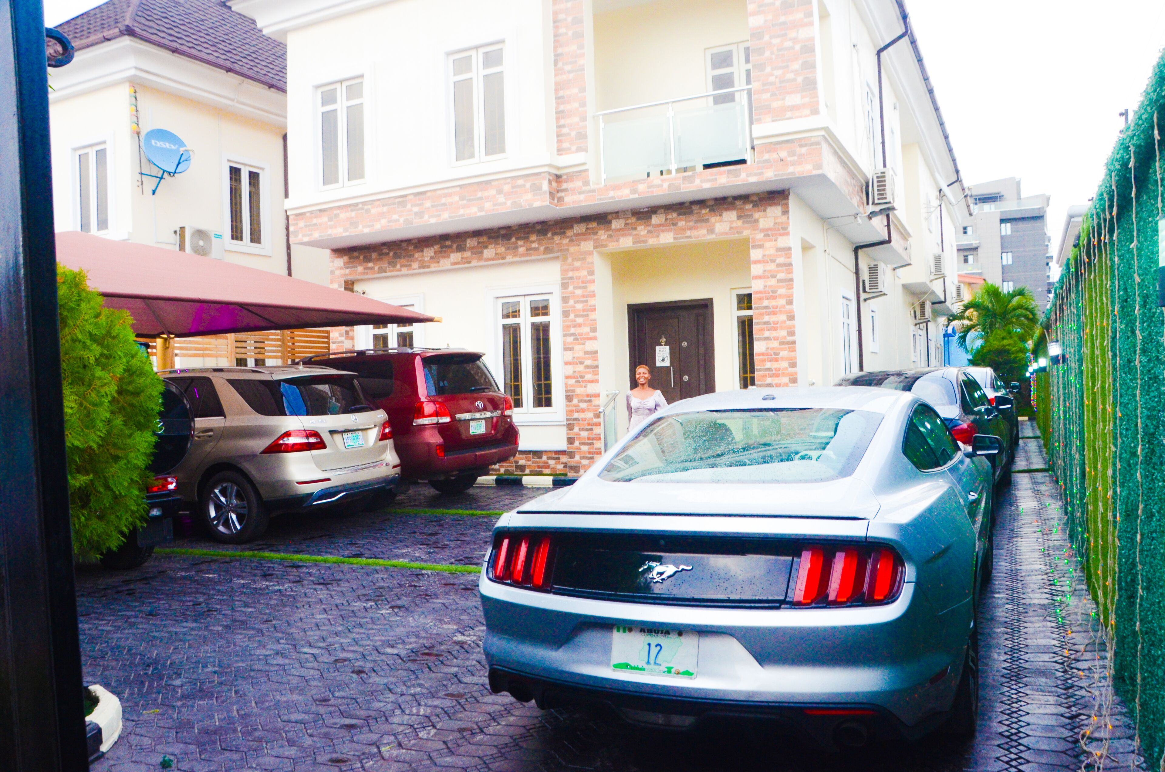 Une rue résidentielle bordée de voitures garées, dont une Mustang argentée, et une maison dotée d’un balcon et d’une antenne parabolique.