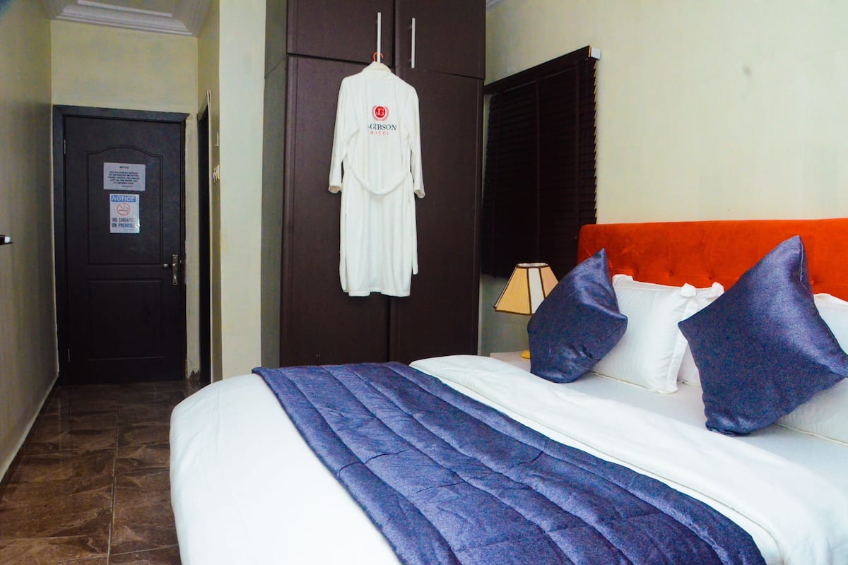 A hotel room with a bed, pillows, a robe hanging on the wall, a door, and a window with blinds.