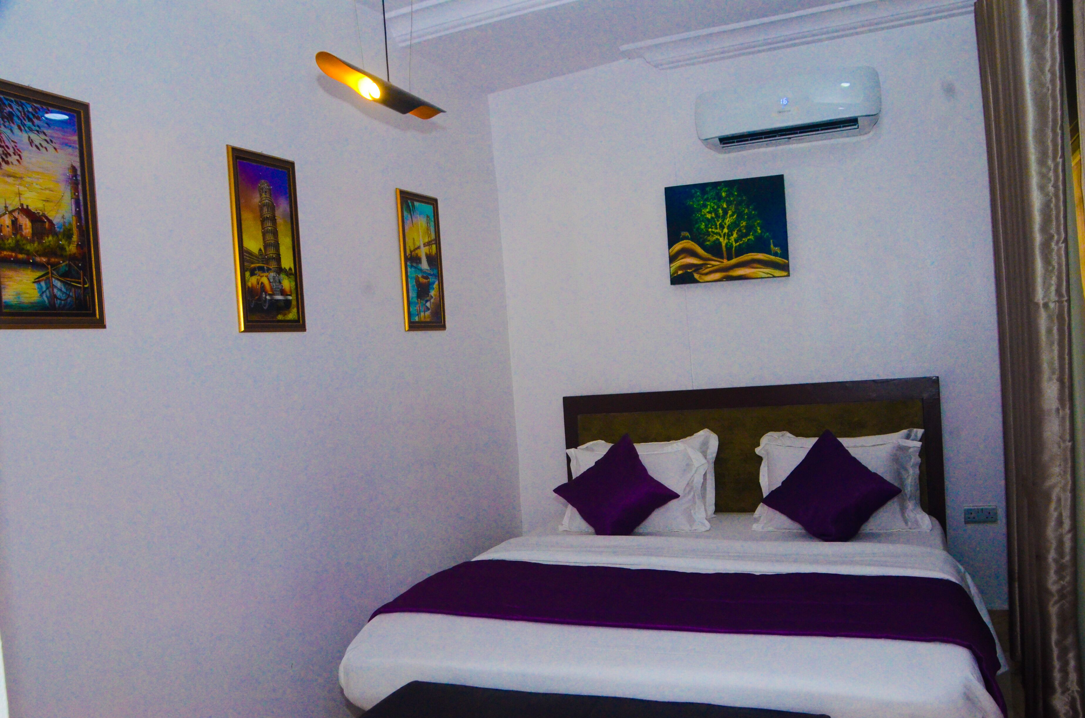 A neatly made bed with a purple blanket and pillows, a ceiling light, and three framed artworks on the wall.