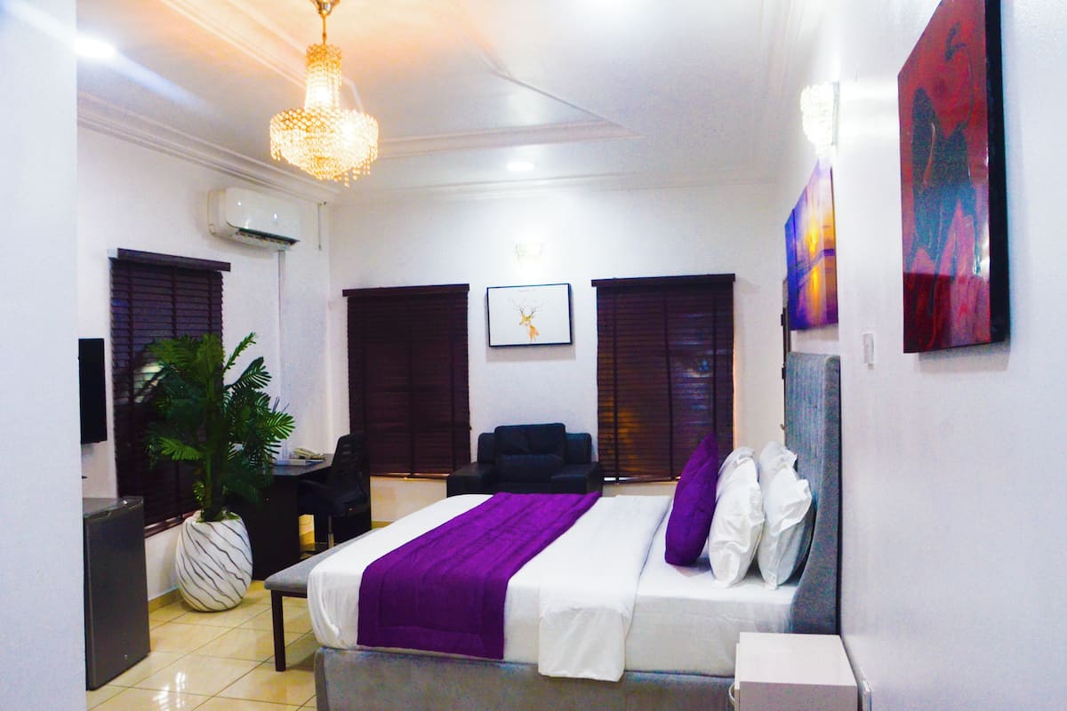 A hotel room with a bed, a desk, a chair, a potted plant, and artwork on the walls.