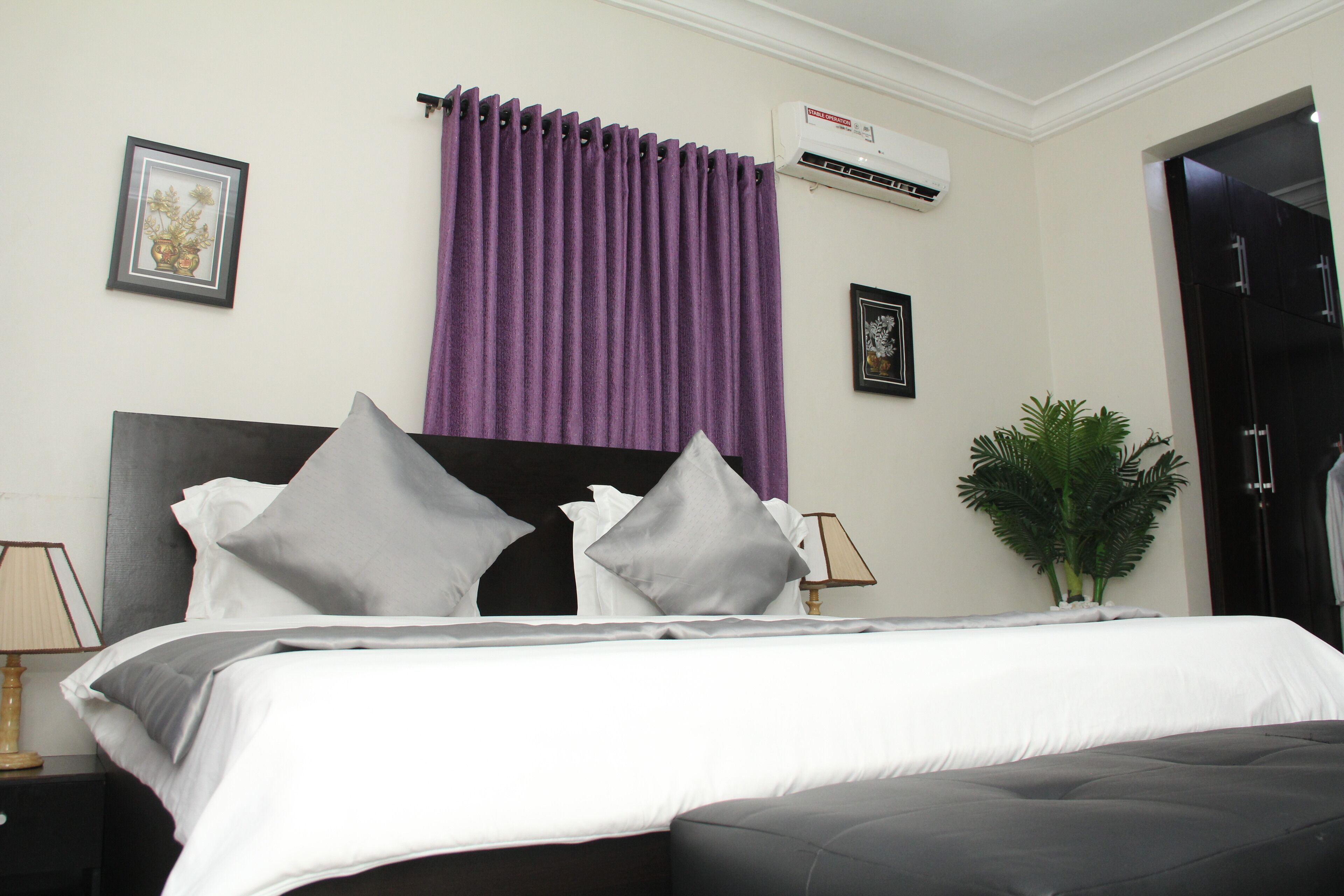 A hotel room with a bed, a nightstand, a lamp, a potted plant, and a door.