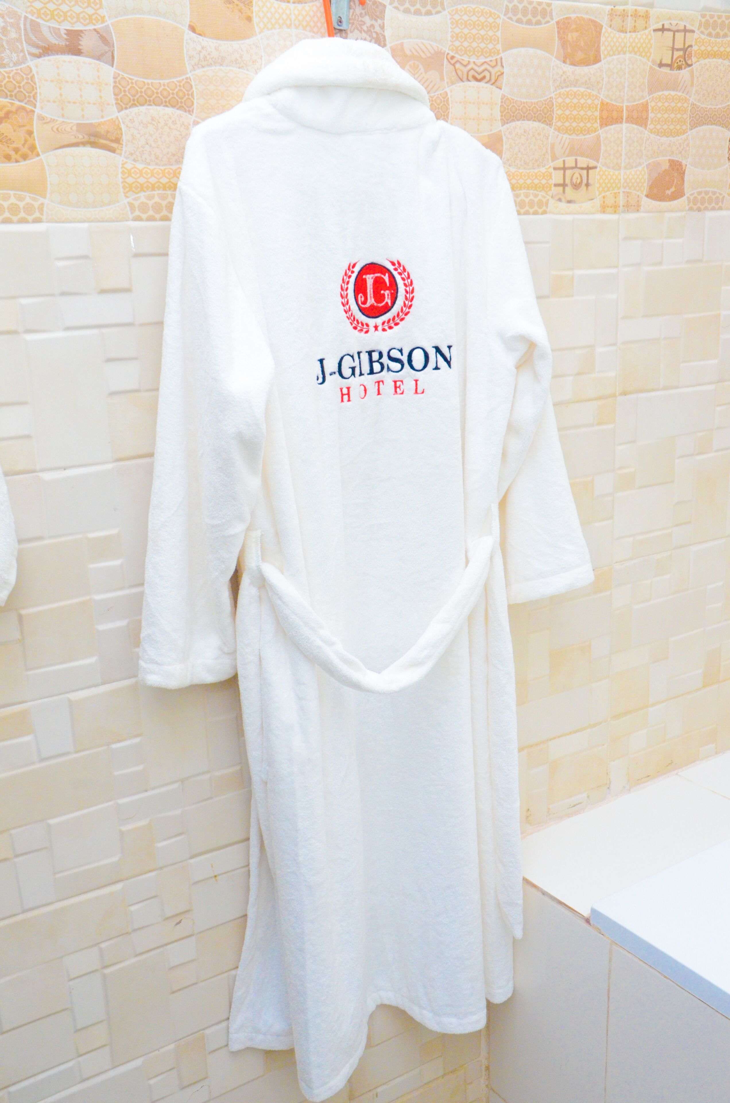 A white robe hanging on a wall with a logo and text that reads "J-Gibson Hotel".