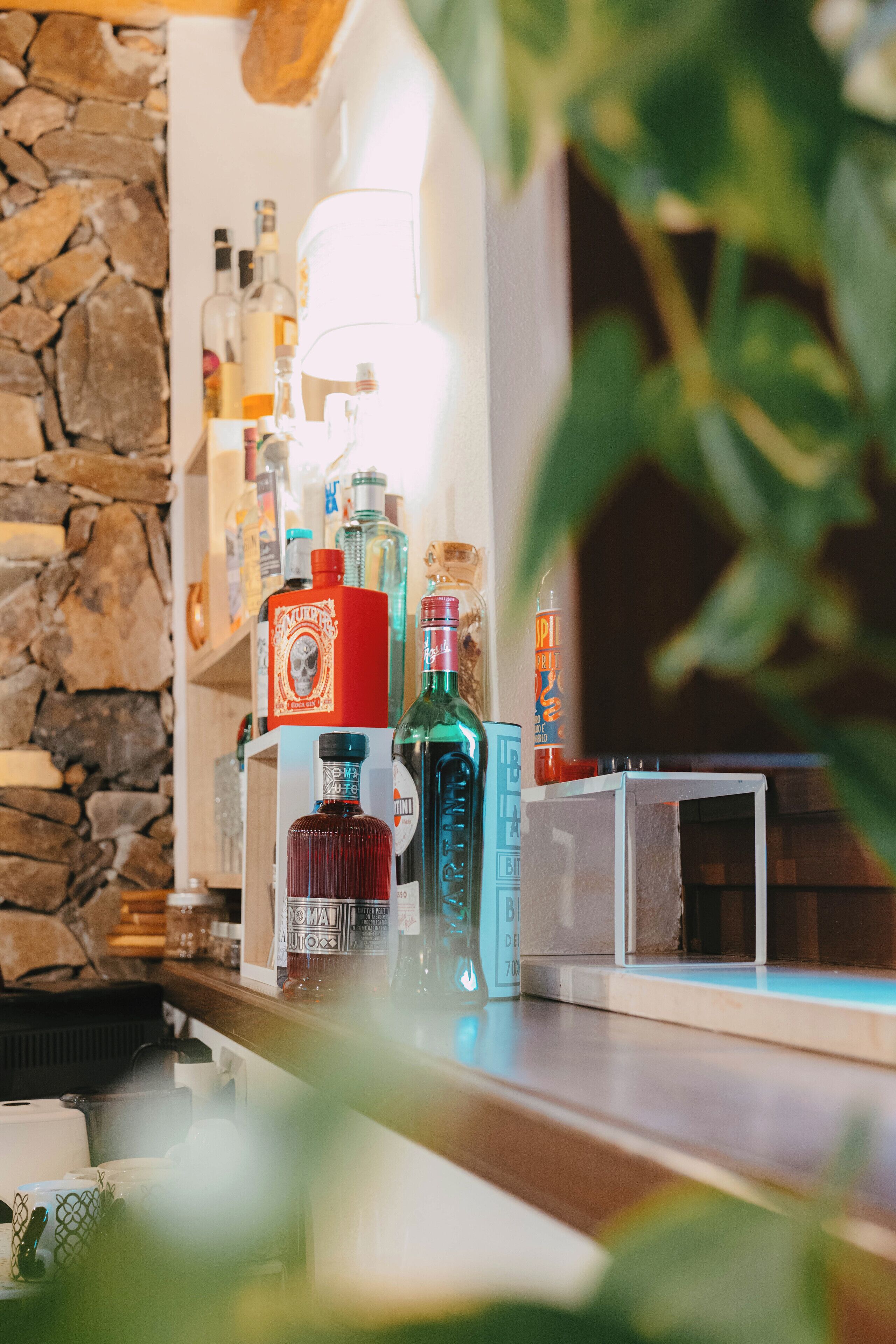 A bar area with various bottles and glasses, a stone wall, and a plant in the foreground.