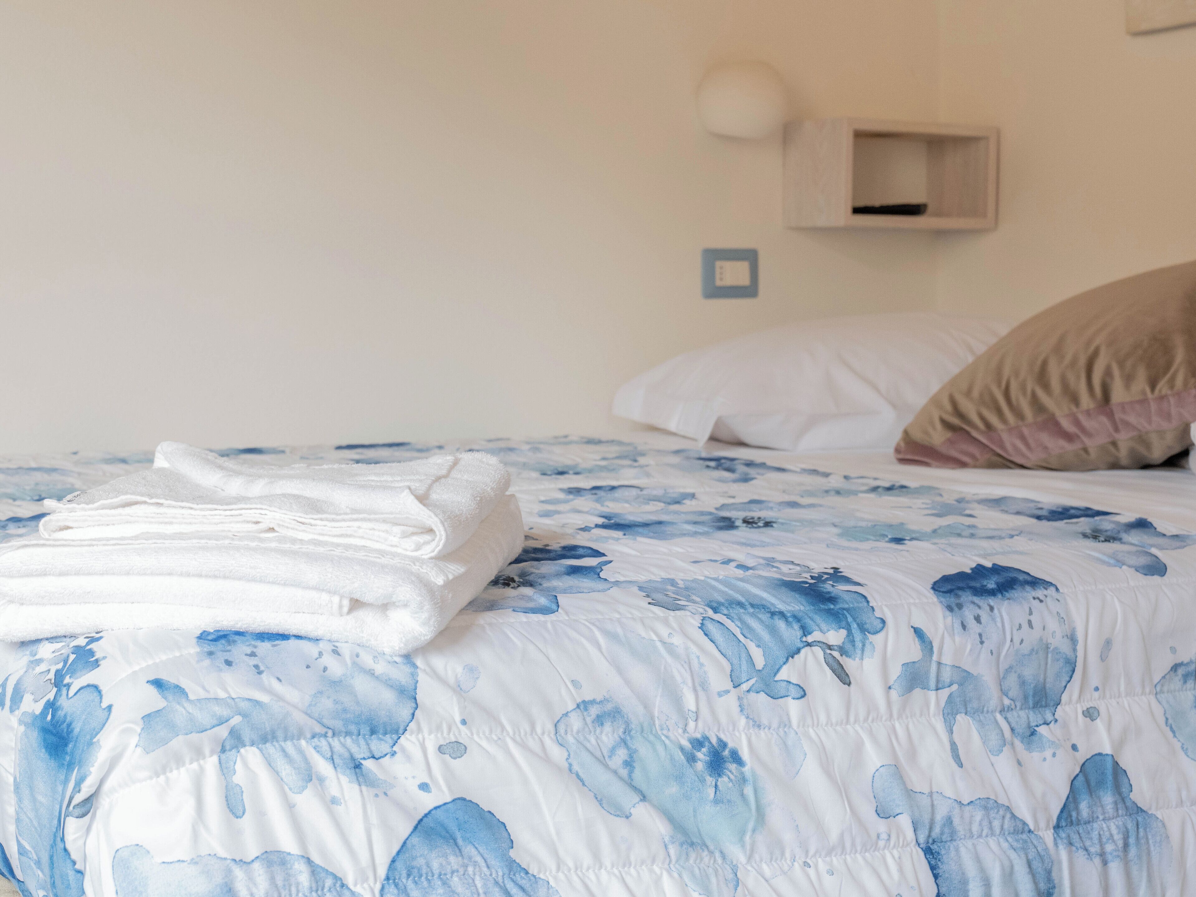 A neatly made bed with a blue floral bedspread, a stack of white towels, and a beige pillow.