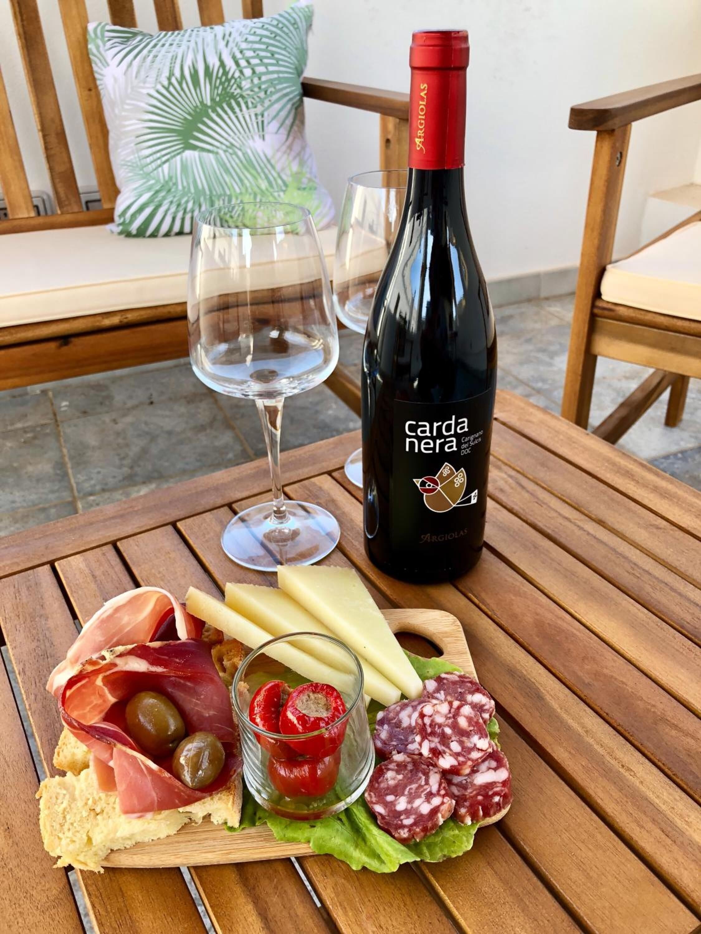 Una mesa de madera con una botella de vino, copas y una tabla de embutidos con carnes, queso, aceitunas y tomates.