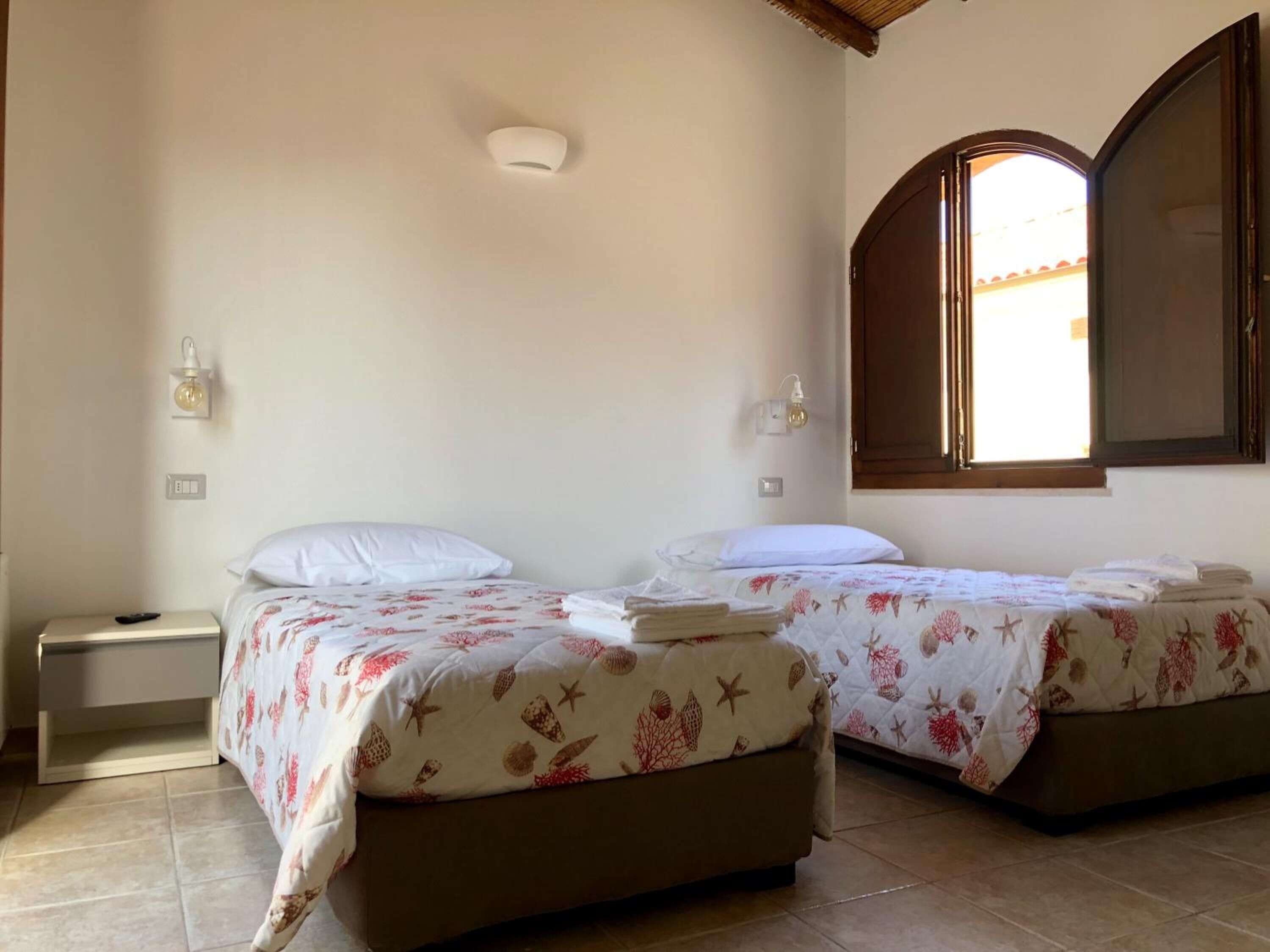 A room with two beds, each with a floral bedspread, a nightstand with a remote control, and a wall-mounted light fixture.
