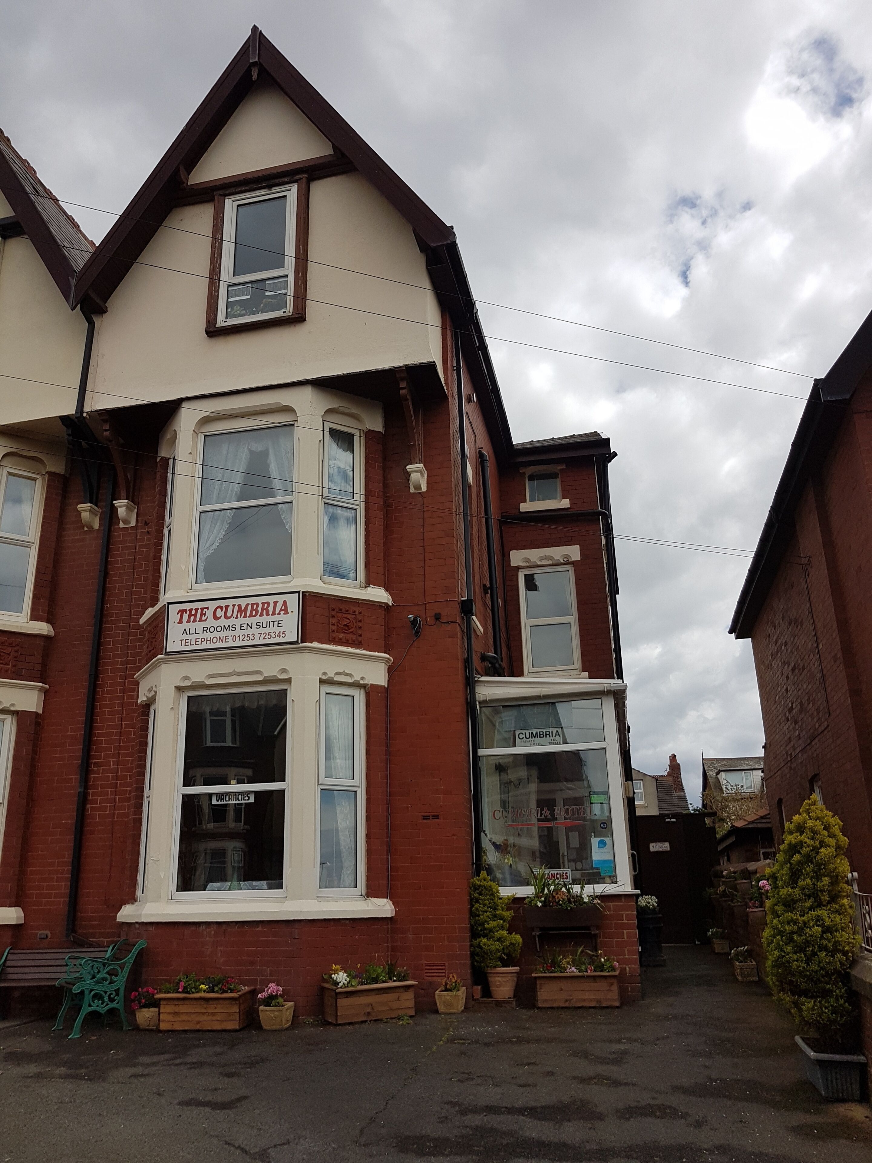 A two-story building with a sign for 'The Cumbria' indicating rooms for rent. There are multiple windows, some with curtains, and the building has a brick facade with a white trim.