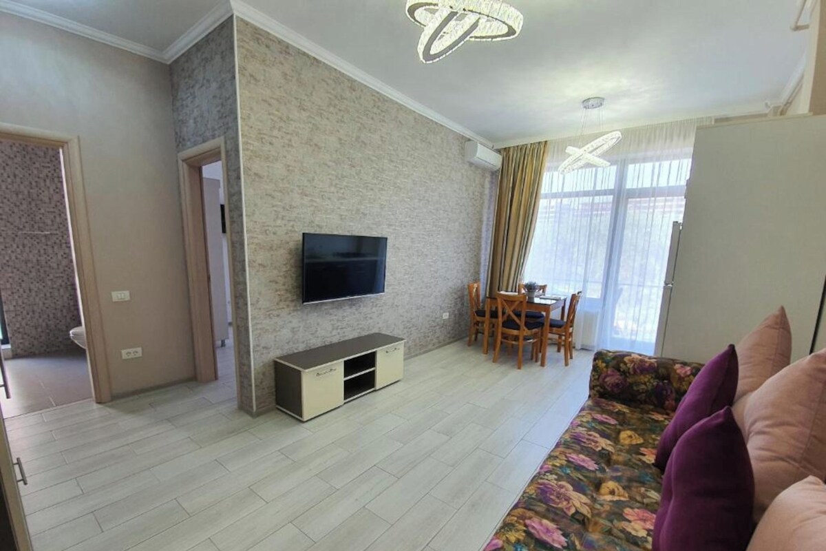 A living room with a flat-screen TV mounted on the wall, a dining area with a table and chairs, and a sofa with floral cushions.