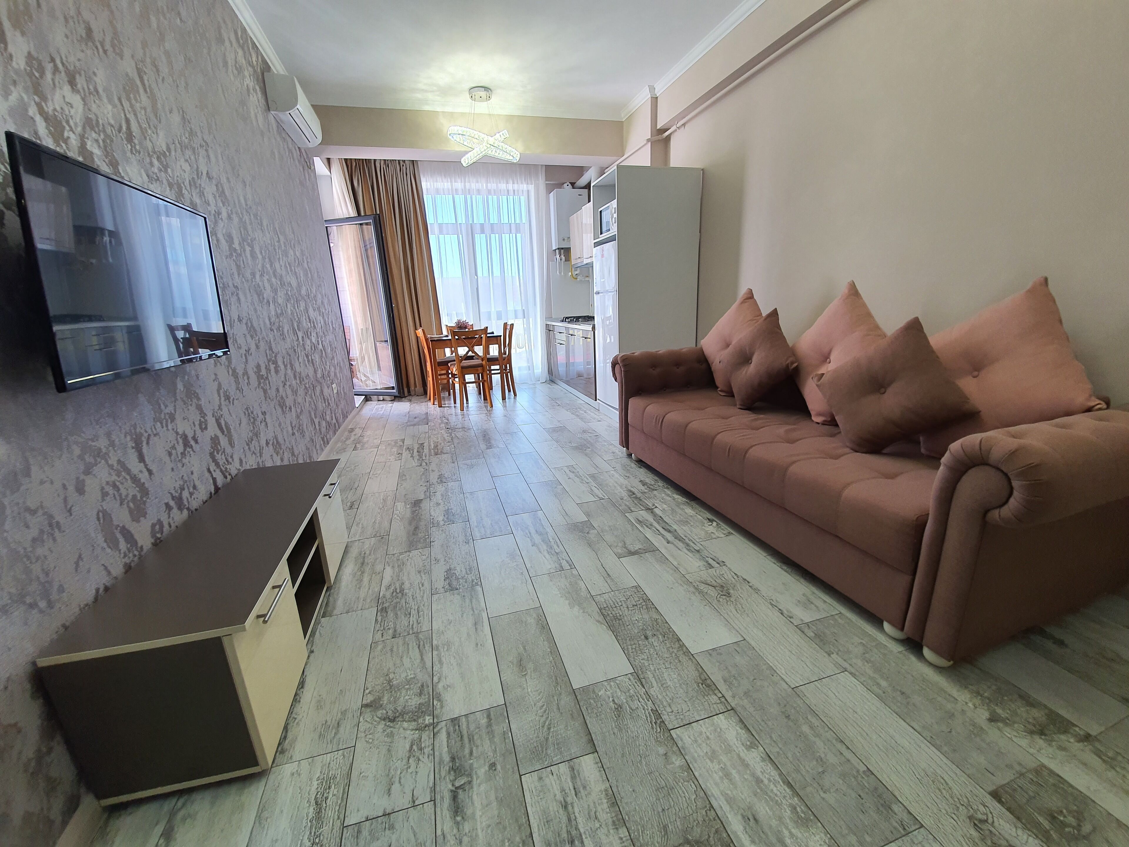 A modern living room with a flat-screen TV, a sofa with cushions, and a dining area with a table and chairs.