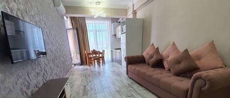 A modern living room with a flat-screen TV, a sofa with cushions, and a dining area with a table and chairs.