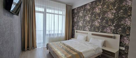 Una habitación de hotel con una cama, cortinas, un papel tapiz floral y un televisor montado en la pared.