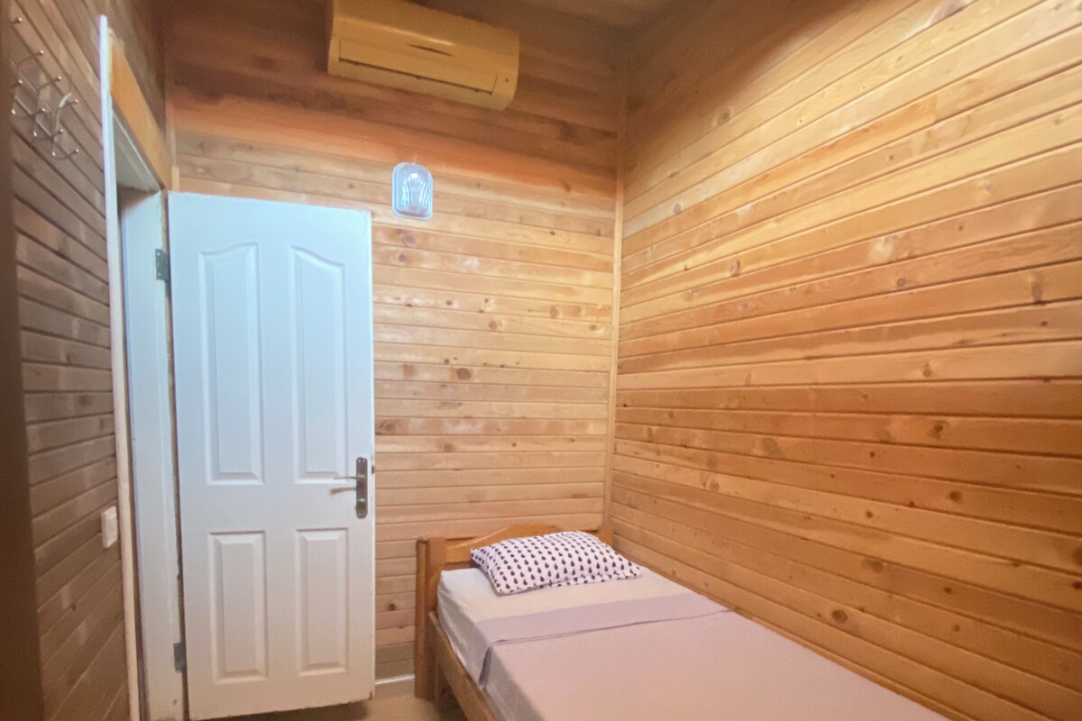 A small, single-bed room with wooden walls and a door leading to another room.