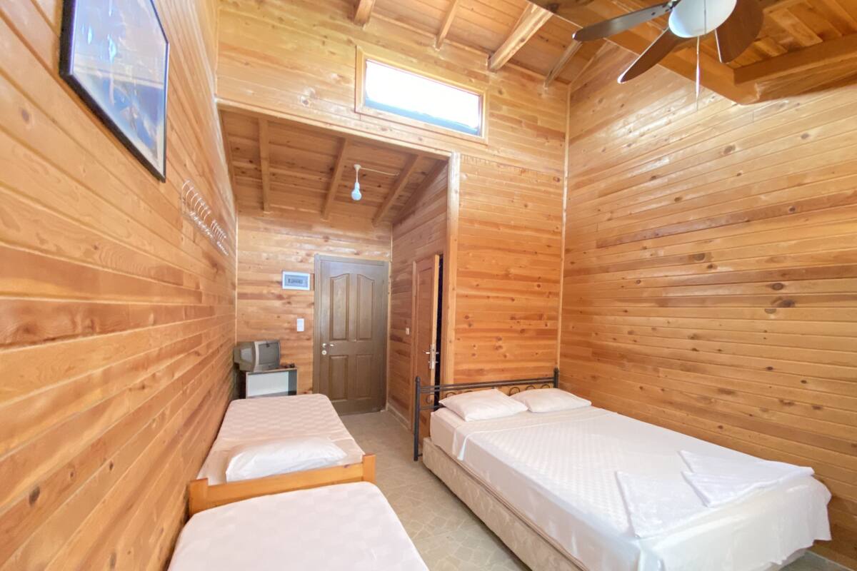 Une chambre de style cabane en bois, avec deux lits simples, un petit bureau avec une chaise, un ventilateur de plafond et une fenêtre.