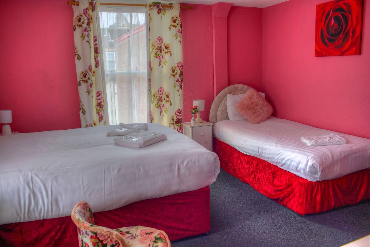 Una habitación con dos camas, una con falda roja y otra con falda blanca. Hay una silla con estampado floral, una mesita de noche con lámpara y un cuadro de una rosa roja en la pared.