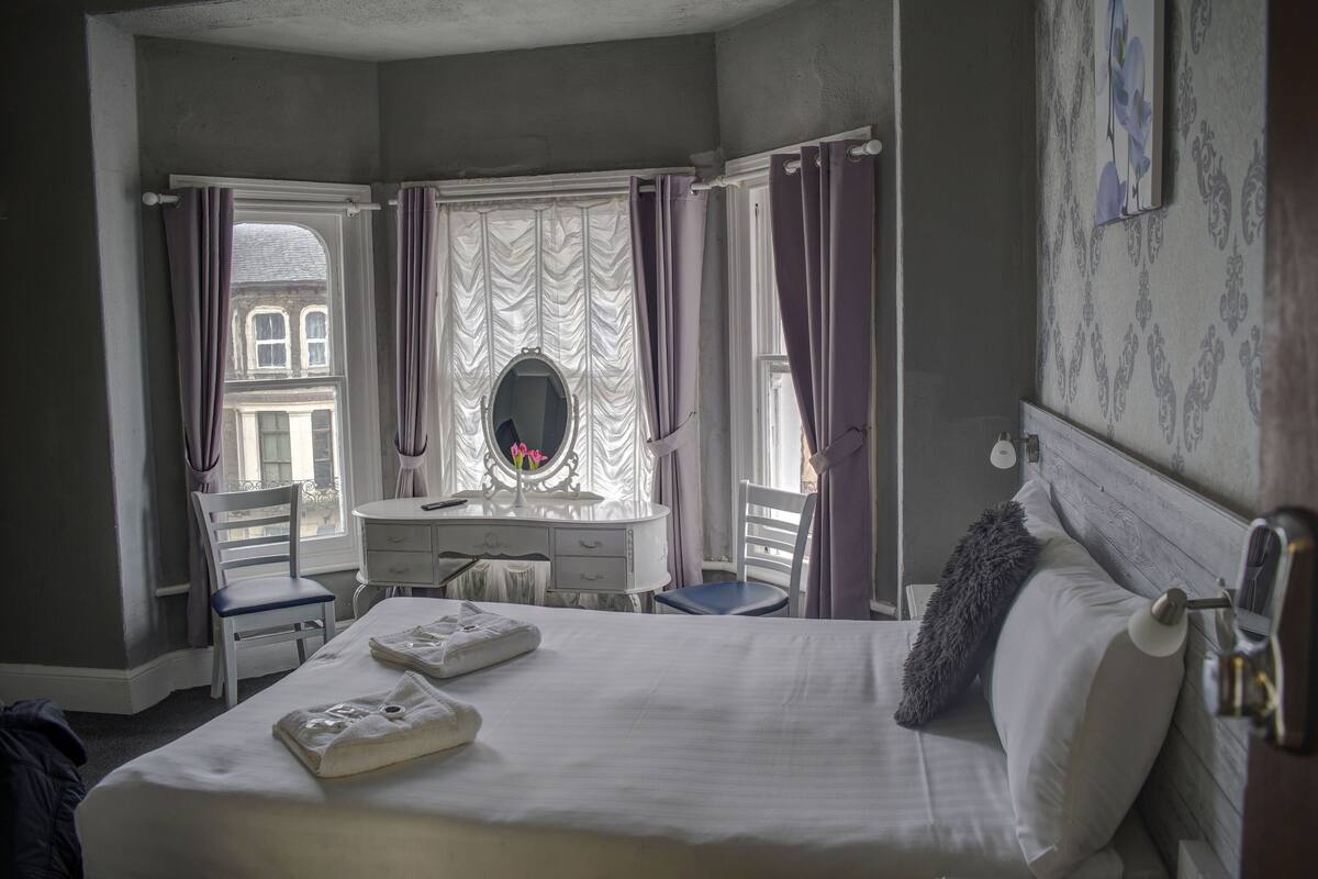 Un dormitorio con cama, cómoda, silla, ventana con cortinas y espejo.