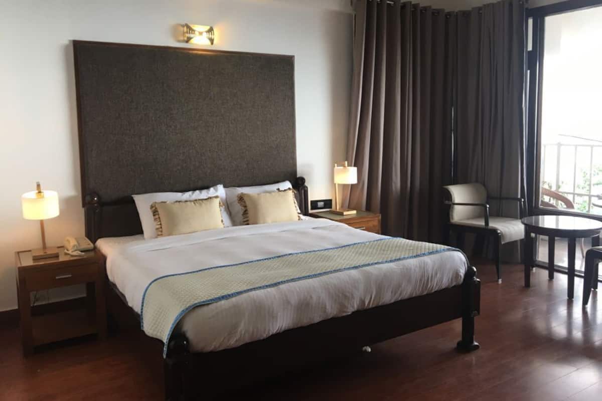 A hotel room with a large bed, bedside lamps, a nightstand, a chair, a table, and a balcony with curtains.