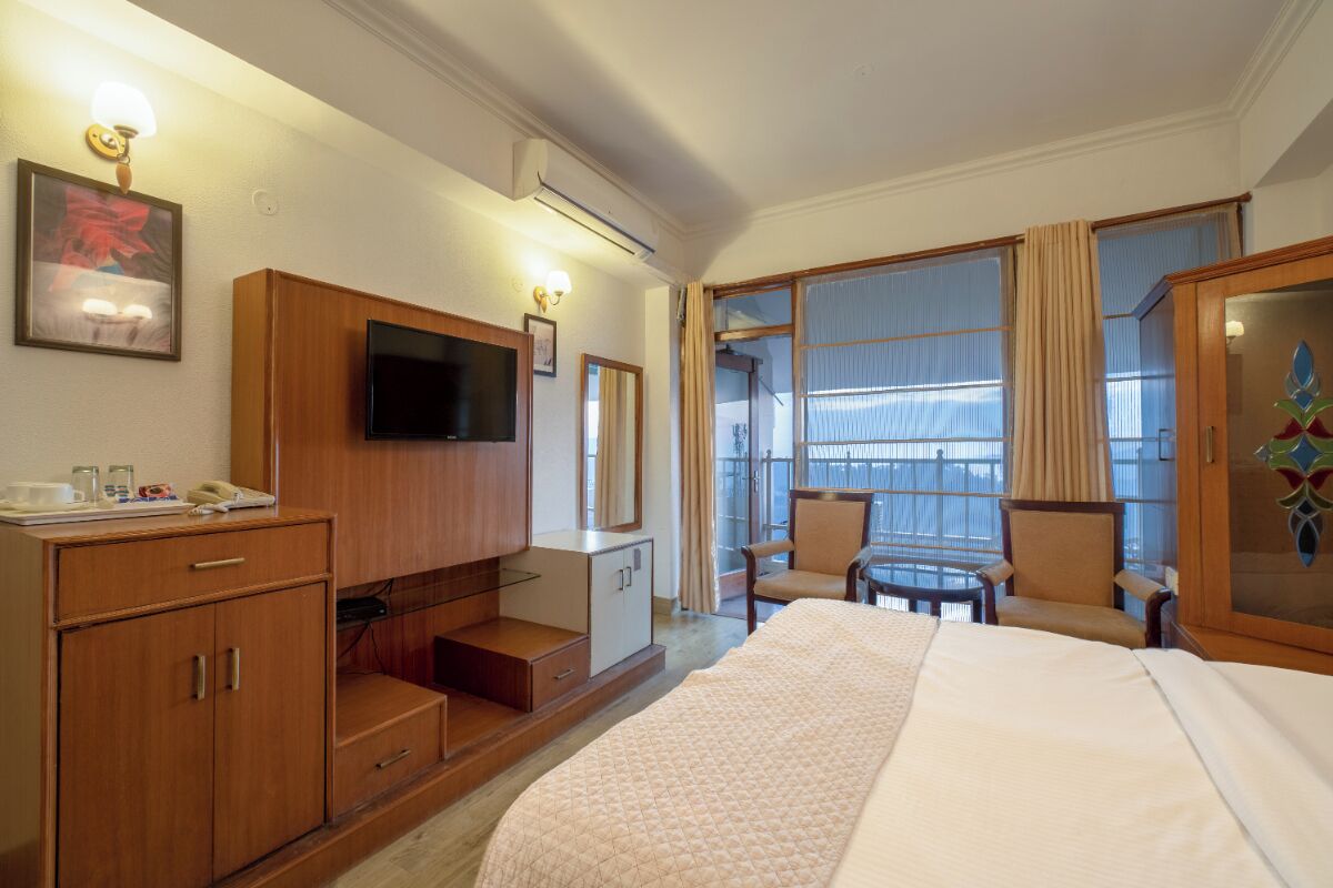 Habitación de hotel con cama, televisión, escritorio con teléfono, silla y ventana con cortinas.