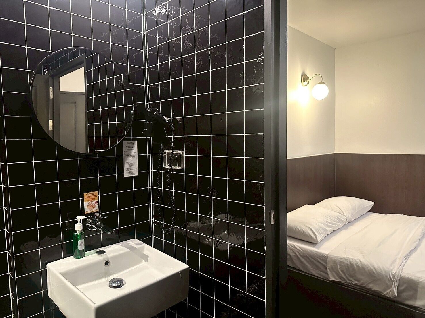 A hotel room with a black-tiled wall, a white sink, a mirror, a bed with white bedding, and a wall-mounted light fixture.