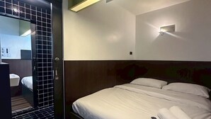 Ένα δωμάτιο ξενοδοχείου με ένα κρεβάτι, ένα κομοδίνο, έναν καθρέφτη και ένα φωτιστικό τοποθετημένο στον τοίχο.