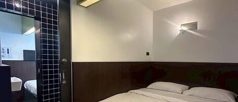 A hotel room with a bed, a bedside table, a mirror, and a wall-mounted light fixture.