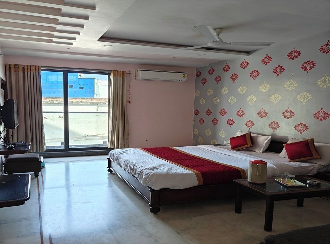 A hotel room with a bed, a desk, a chair, a table, a ceiling fan, and a large window.