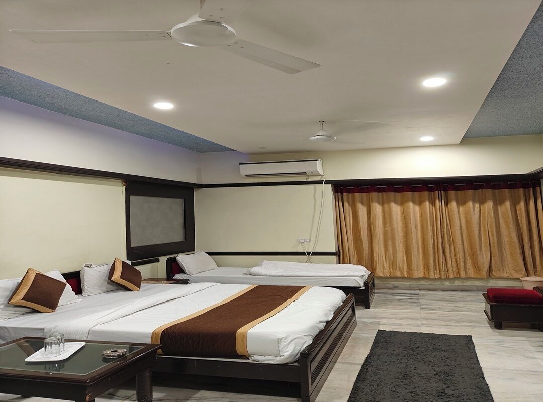 A hotel room with two beds, a desk with a glass top, a ceiling fan, an air conditioning unit, and a window with curtains.