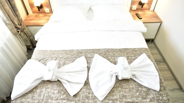 Quarto de hotel com cama arrumada, duas mesas de cabeceira com abajures e uma janela grande com cortinas.