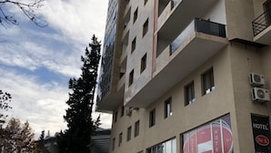 Um hotel com vários andares, varandas, um letreiro visível para 'The K Hotel' e uma loja com a inscrição 'ALCORIUM'.