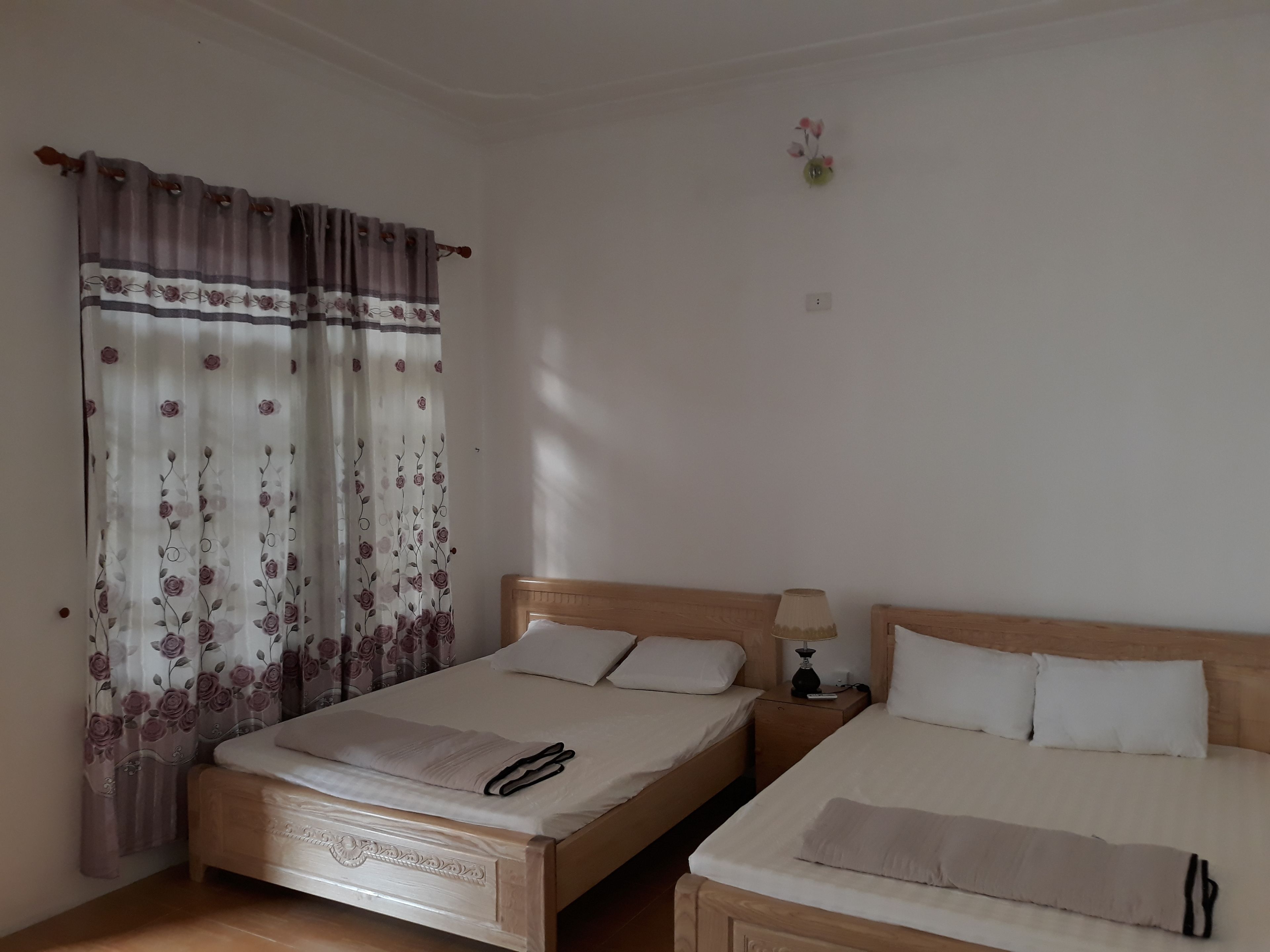 Une chambre avec deux lits simples, une tête de lit en bois, une table de chevet avec une lampe et un rideau à motifs floraux.
