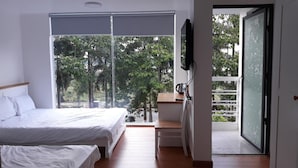 A bedroom with a bed, a desk, and a TV. There is a balcony with a view of trees.