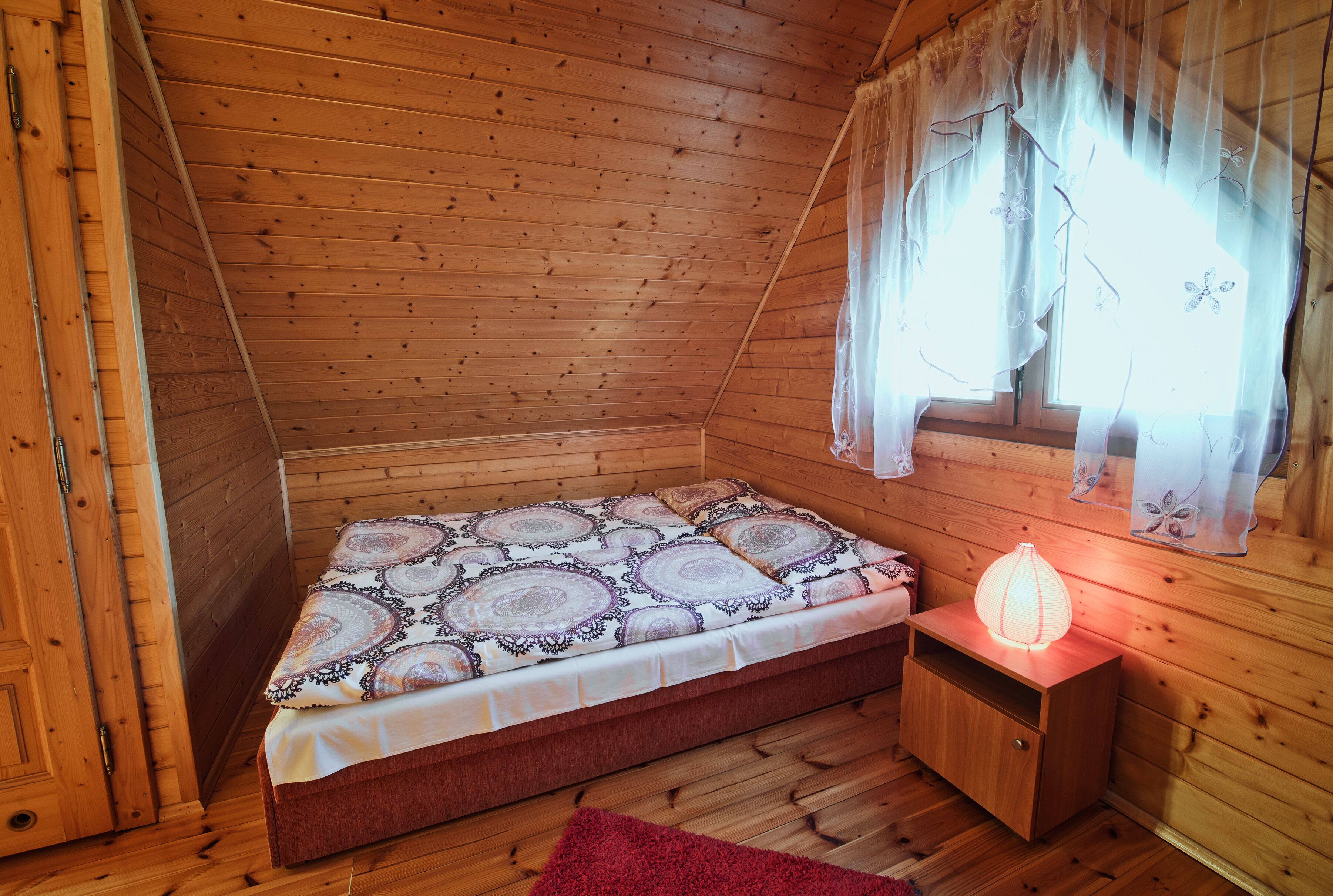 A wooden cabin room with a bed, a nightstand, and a window with curtains.