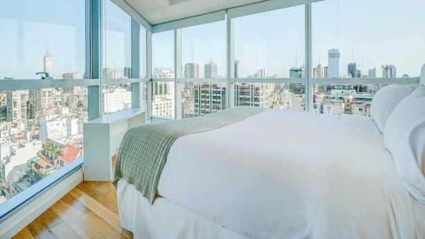 A bedroom with a large bed, a view of a cityscape through floor-to-ceiling windows, and a wooden floor. - Nido x Bellini Esmeralda (Buenos Aires)