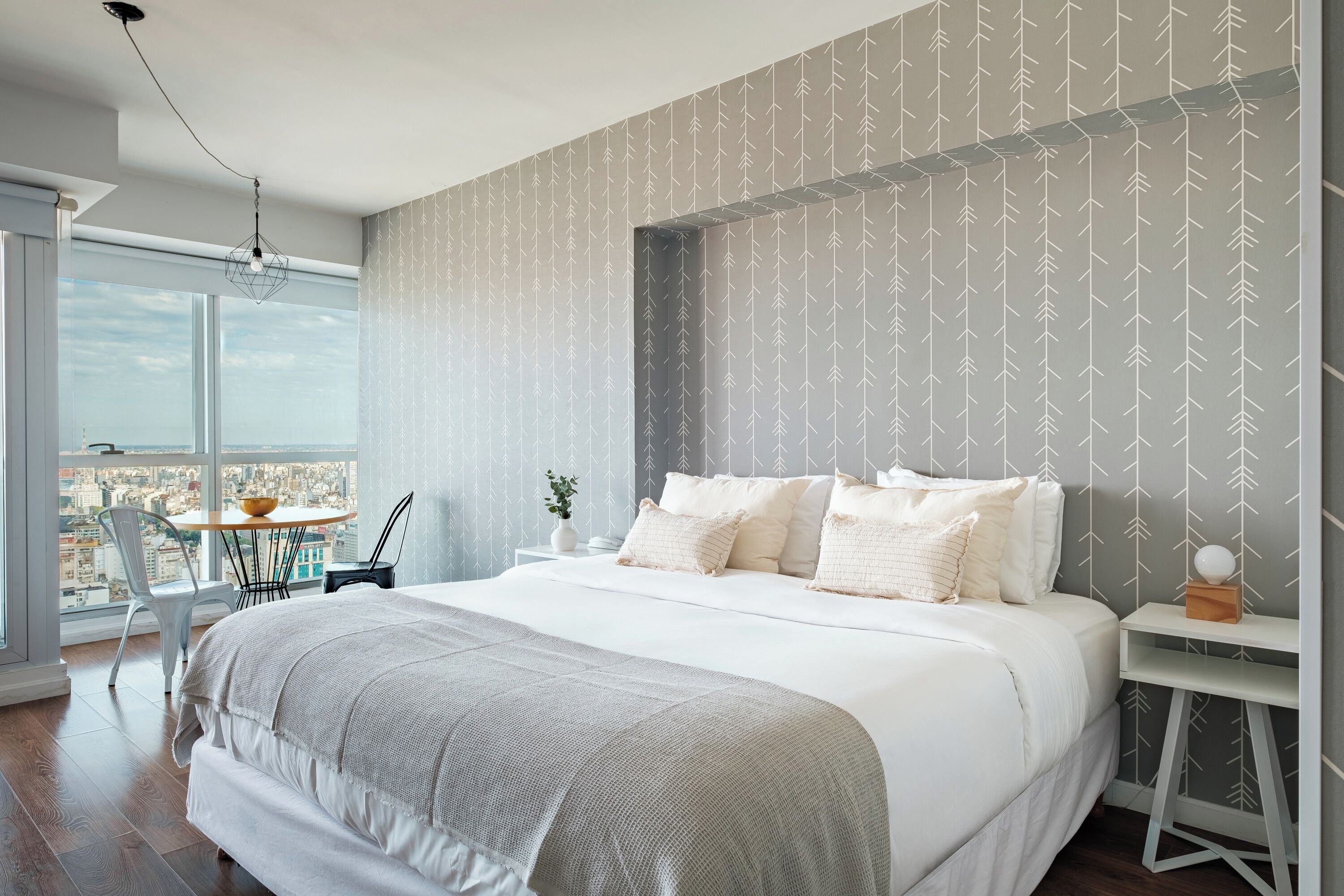 A modern bedroom with a large bed, a view of the city, and a balcony with a table and chairs.