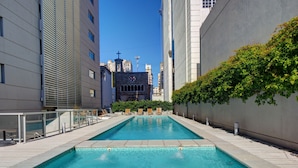 A rectangular swimming pool with clear blue water, surrounded by modern buildings, a visible church tower, and a lush green hedge. - Nido x Bellini Esmeralda (Buenos Aires)