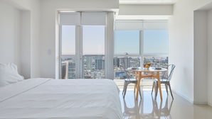 A modern hotel room with a large bed, a dining table with chairs, and a city view through large windows. - Nido x Bellini Esmeralda (Buenos Aires)