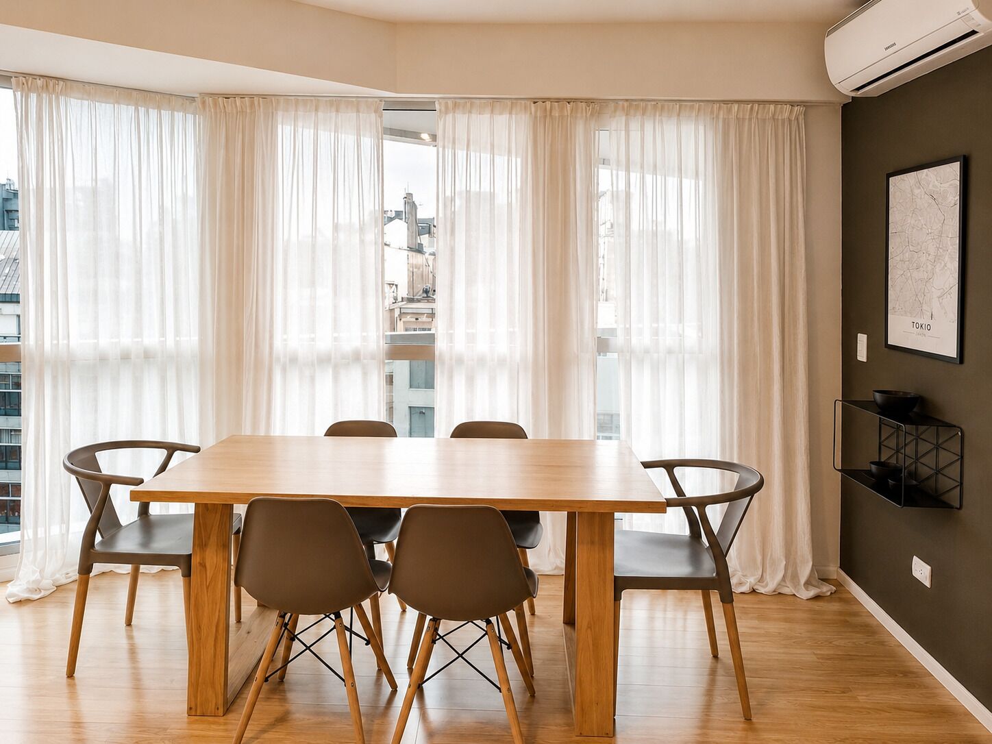 A modern dining area with a wooden table and chairs, large windows with sheer curtains, and a cityscape view.
