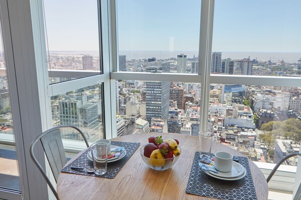 A table with a fruit bowl, a cup, and a glass, overlooking a cityscape with buildings and a view of the ocean.