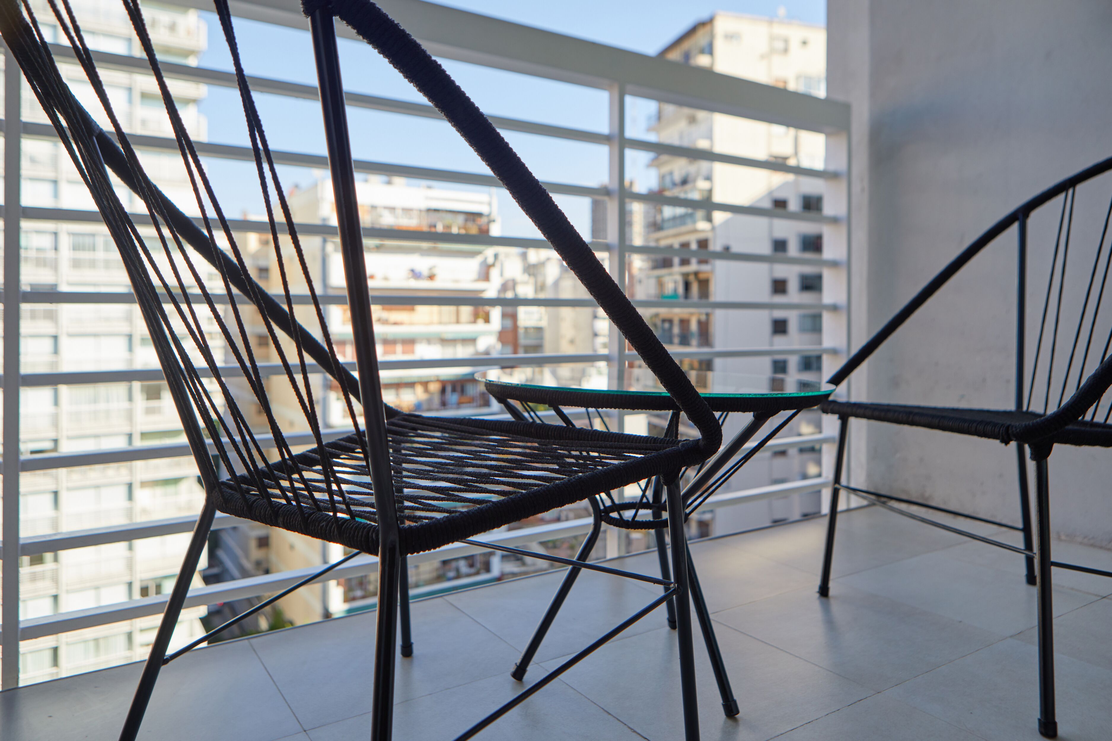 A balcony with two wicker chairs and a small table, overlooking a cityscape with multiple buildings.