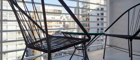 A balcony with two wicker chairs and a small table, overlooking a cityscape with multiple buildings.