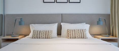 Un lit bien fait, avec du linge de lit blanc et deux coussins décoratifs, flanqué de deux tables de chevet surmontées de lampes, dans une chambre d’hôtel moderne.